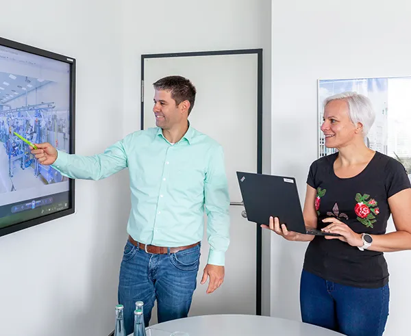 Zwei LTS-Mitarbeitende stehen in einem Büro; der Mann zeigt auf einen Wand-Bildschirm mit technischen Zeichnungen, die Frau hält ein Tablet.