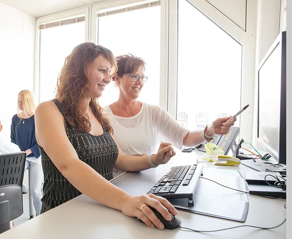 Zwei Frauen arbeiten gemeinsam an einem Computer in einem hellen Büro mit großen Fenstern.